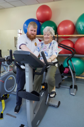 Female patient on an exercise bike being assisted by a musculoskeletal practitioner.