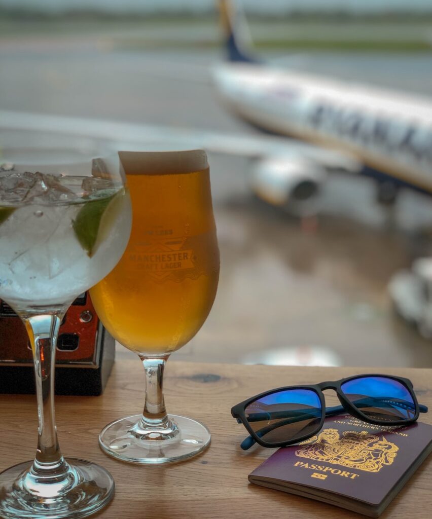 blue sunglasses on a table with a British passport and two alcholic drinks.  Part of an aeroplane can be seen in the background through an airport window.