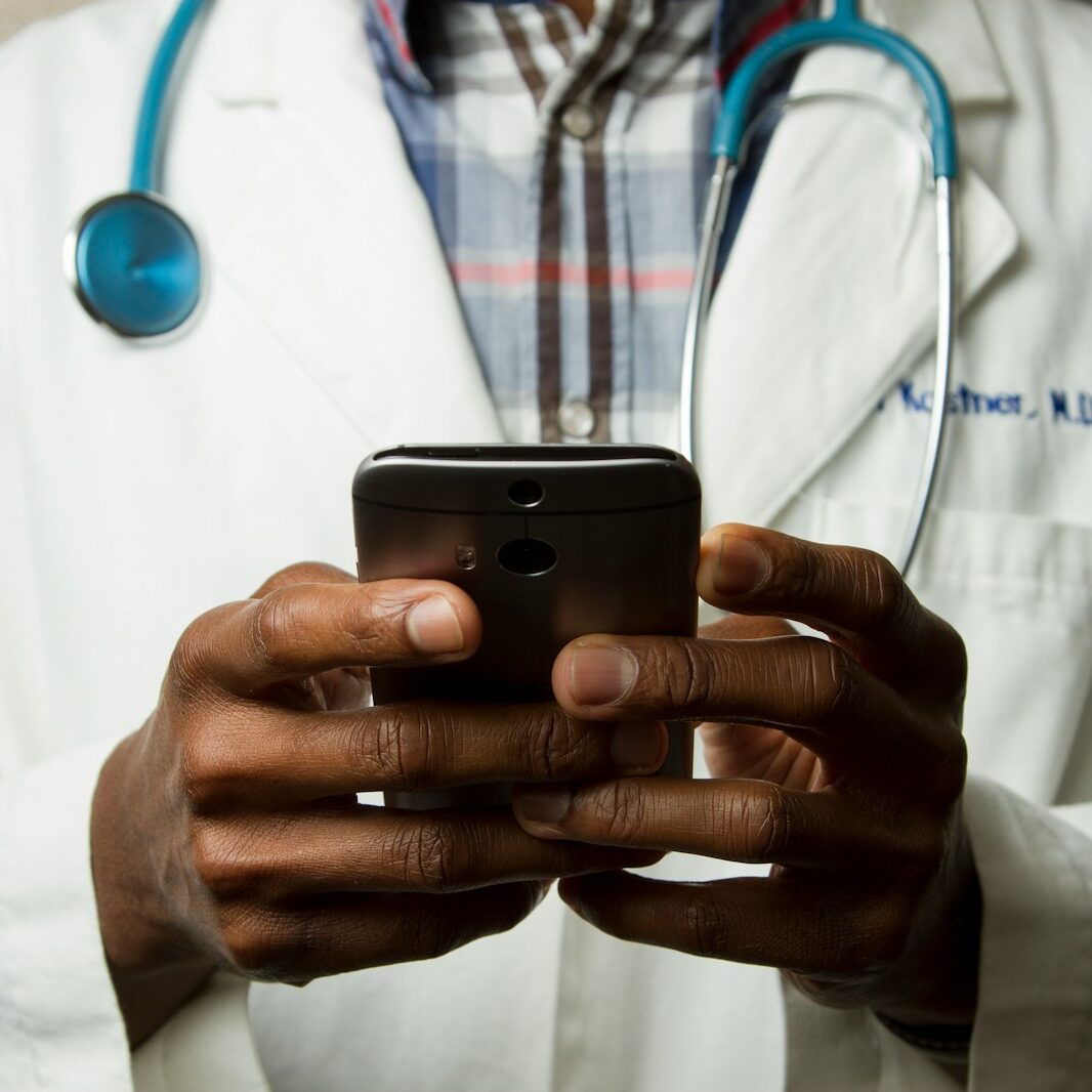 person wearing laboratory gown with green stethoscope on neck using phone while standing